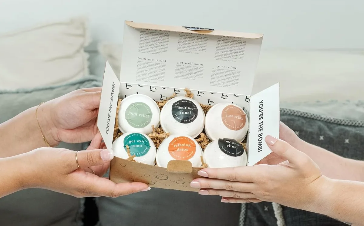 Hands holding an open gift box of round bath bombs with labeled scents