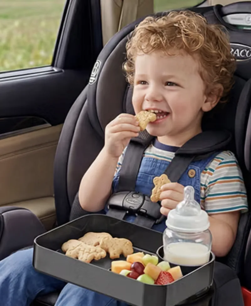 Child in a car seat with a snack tray holding cookies, fruit and a bottle