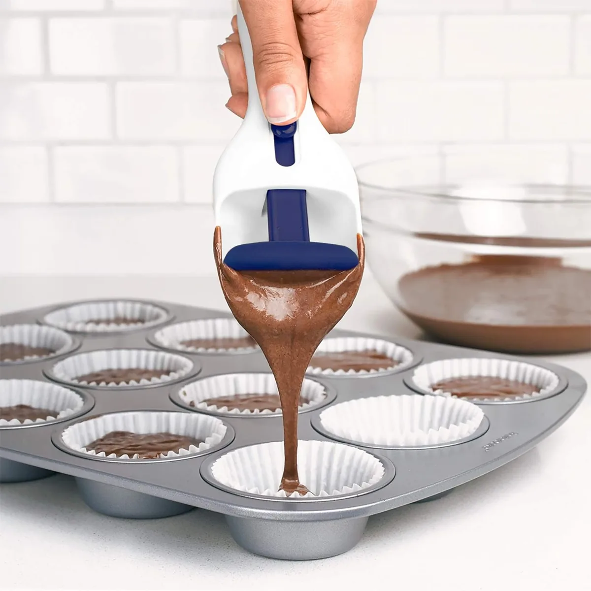 Hand using a batter scoop to dispense cupcake batter into a lined muffin tin