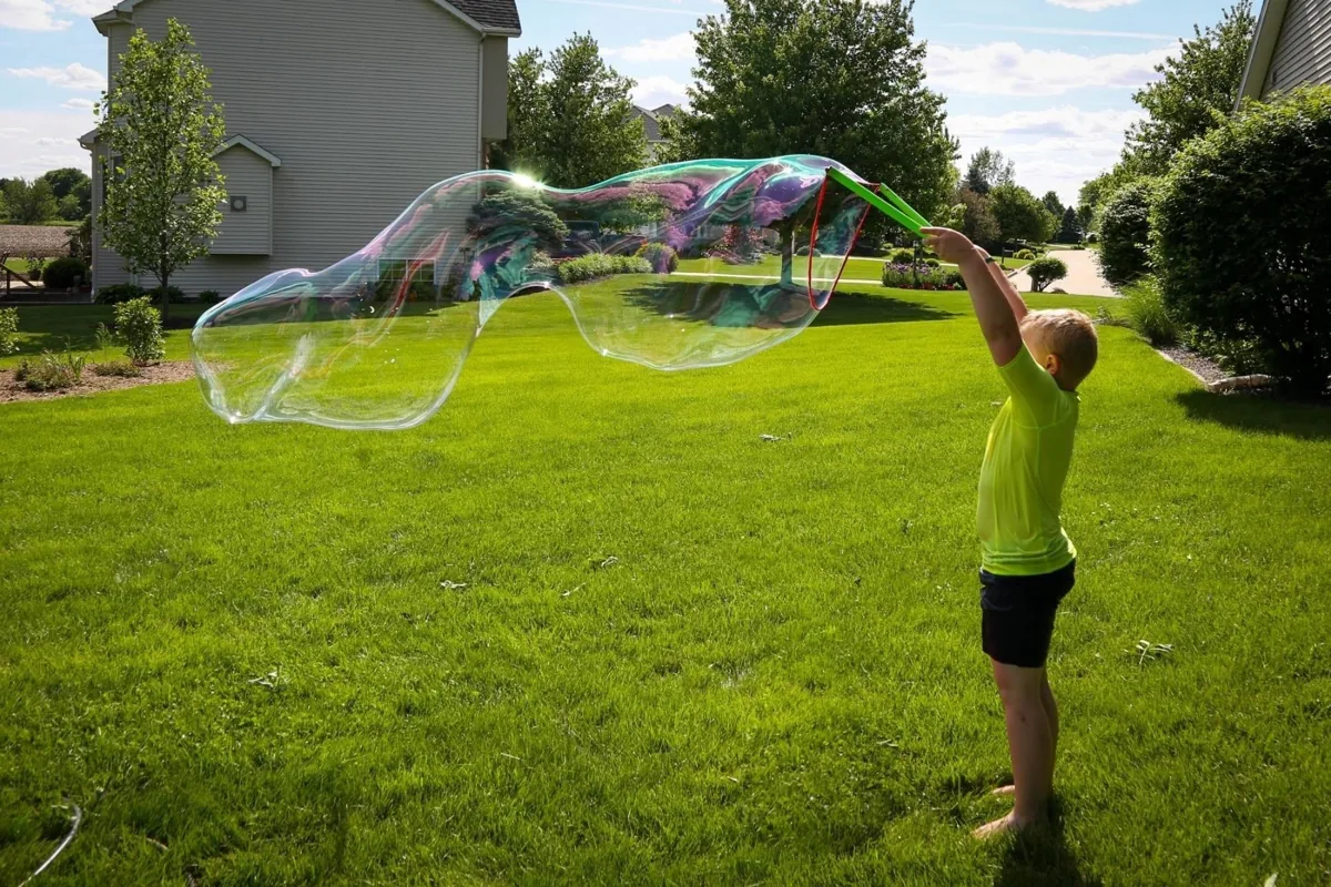 Child making a giant bubble with a two-handle wand in a backyard