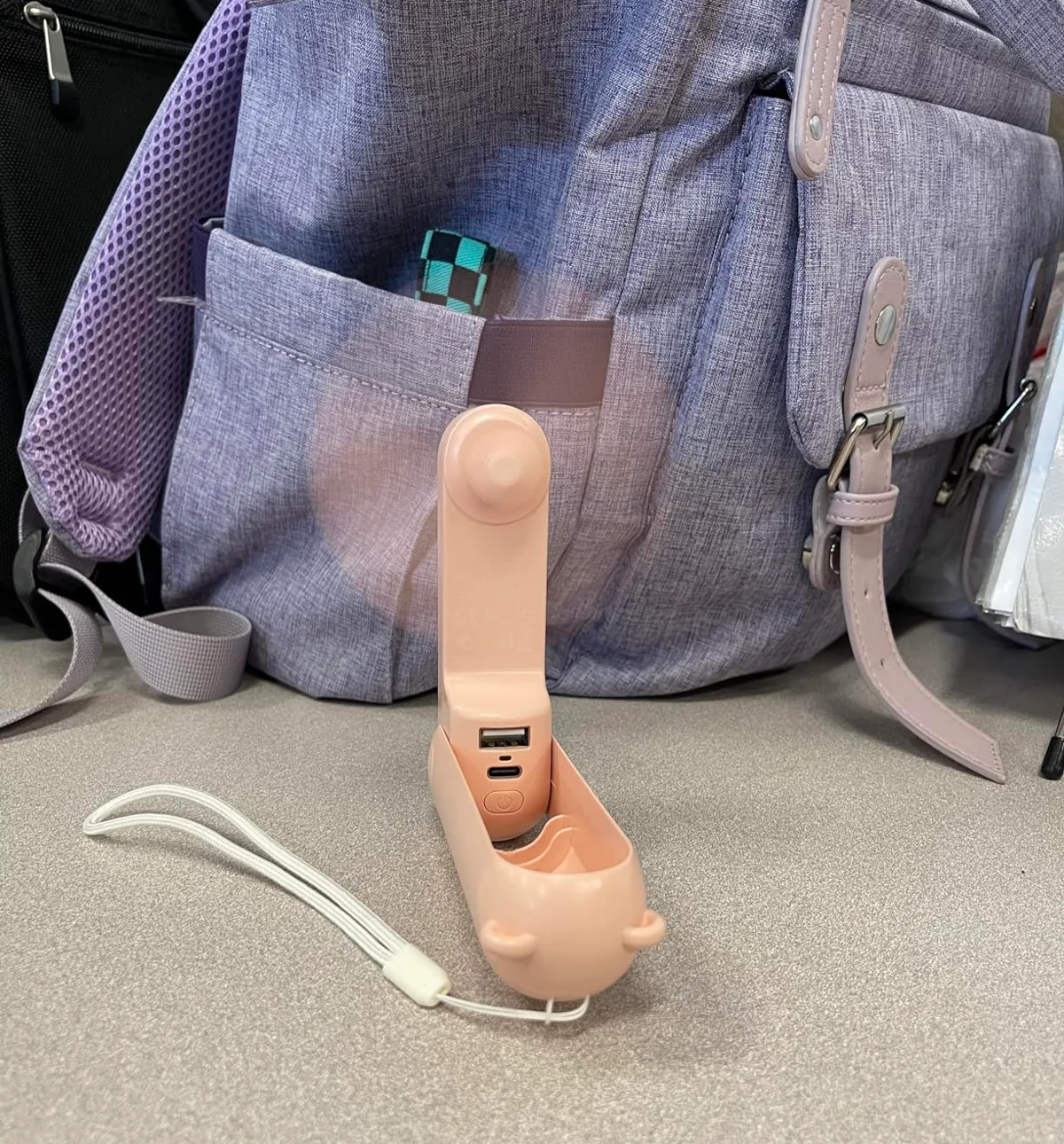 Pink JISULIFE mini fan folded upright next to a backpack