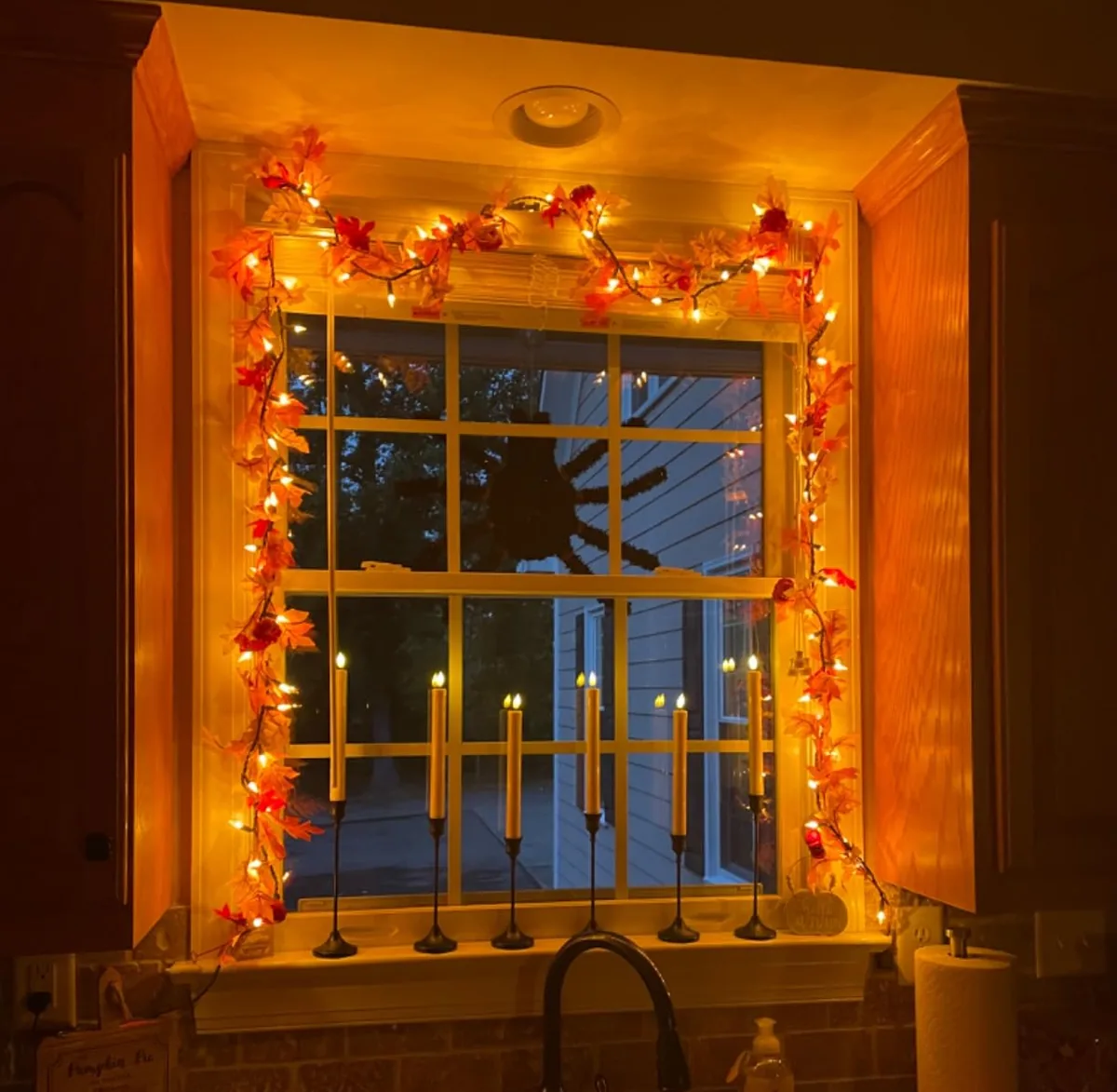 Kitchen window framed with a lighted orange maple leaf garland and candles