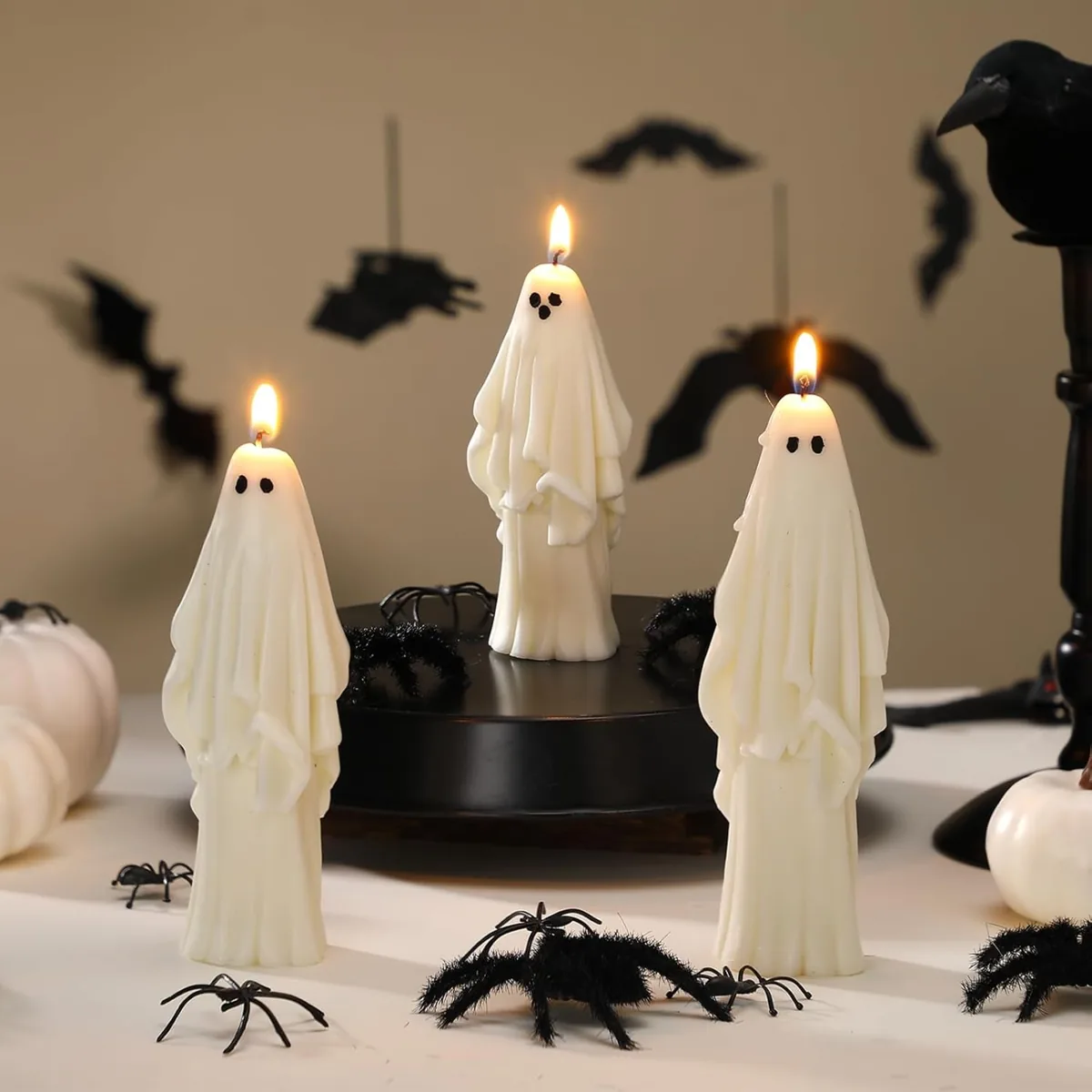 Three lit ghost-shaped candles on a table with toy spiders and paper bats