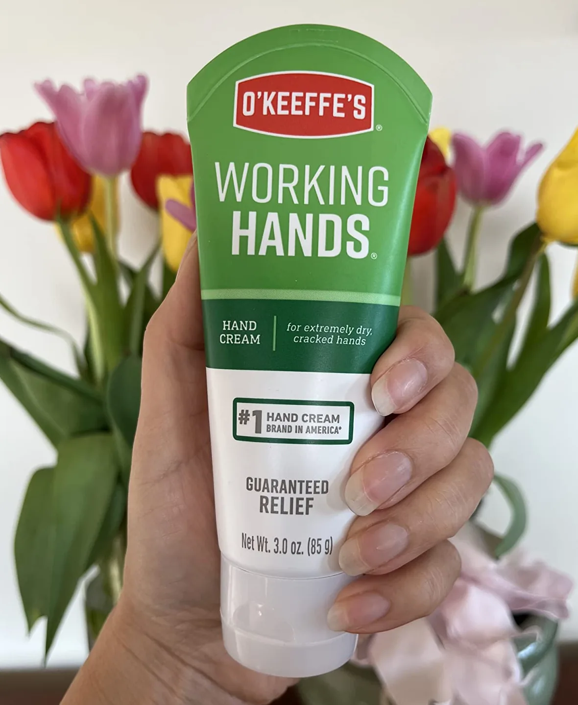 Hand holding a tube of O'Keeffe's Working Hands hand cream in front of tulips