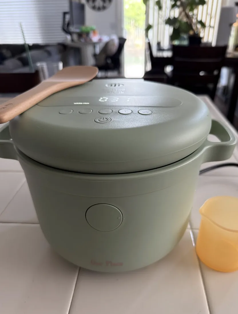 Sage green Our Place rice cooker with a wooden paddle and measuring cup