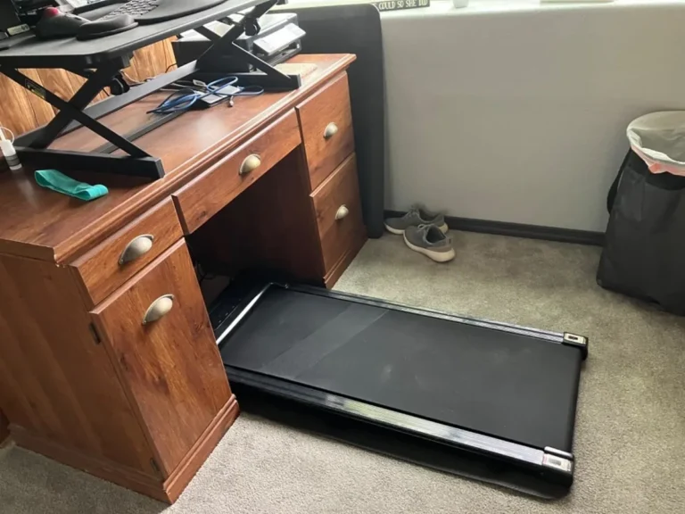 Black portable walking pad slid partly under a wooden home office desk