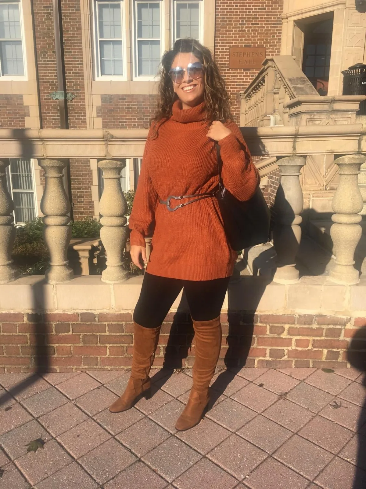 Rust turtleneck sweater dress cinched with a belt over black leggings and tall suede boots